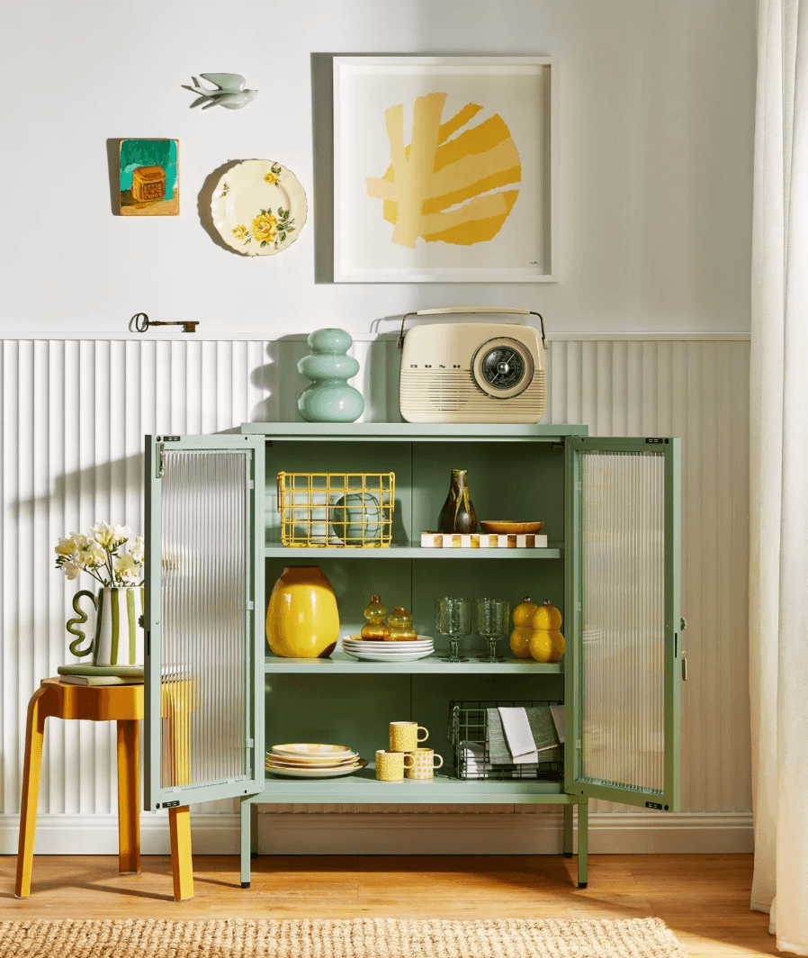 A sage green open cabinet with glass doors stands in a styled room setting, styled with dishes, a vintage radio, and decorative items. The room features a white wainscoted wall, a framed yellow abstract print, and a small wooden stool with a vase of flowers.