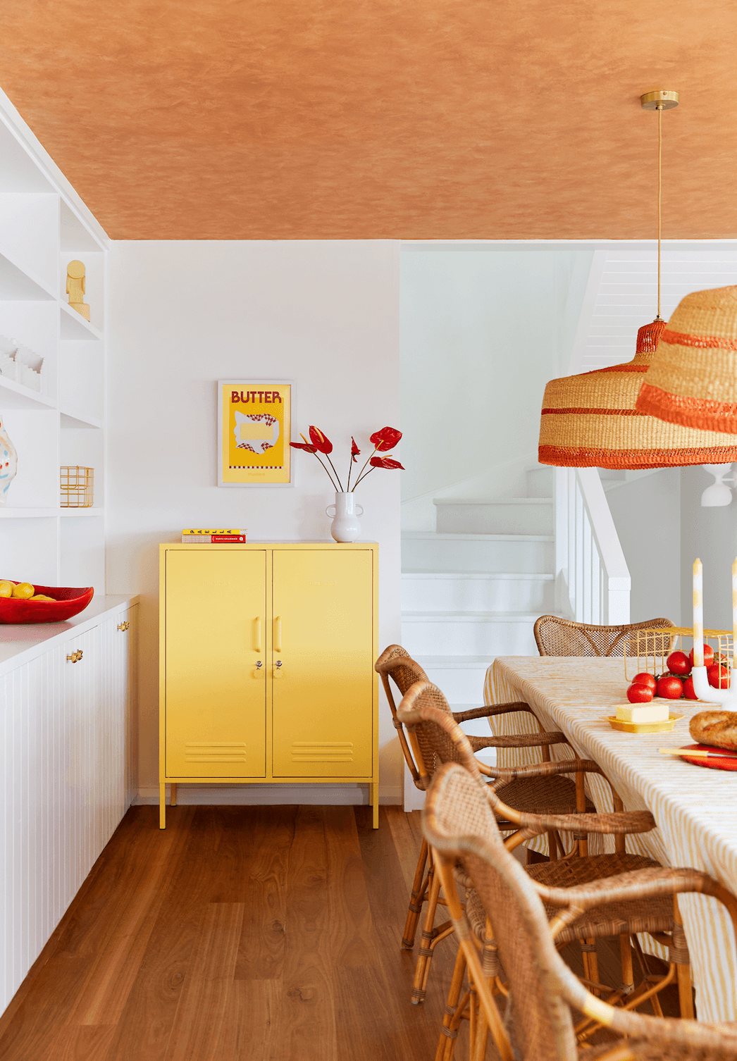 A bright yellow metal cabinet with double doors stands in a modern kitchen or dining space with a wood floor, white walls, a striped pendant light, and woven dining chairs.