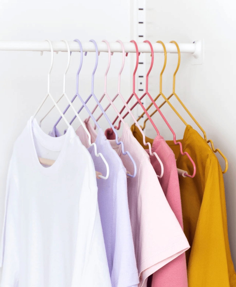 A set of pastel-colored clothes hangers in white, lavender, pink, red, and mustard yellow, holding a row of matching light-colored tops on a white wall rack.