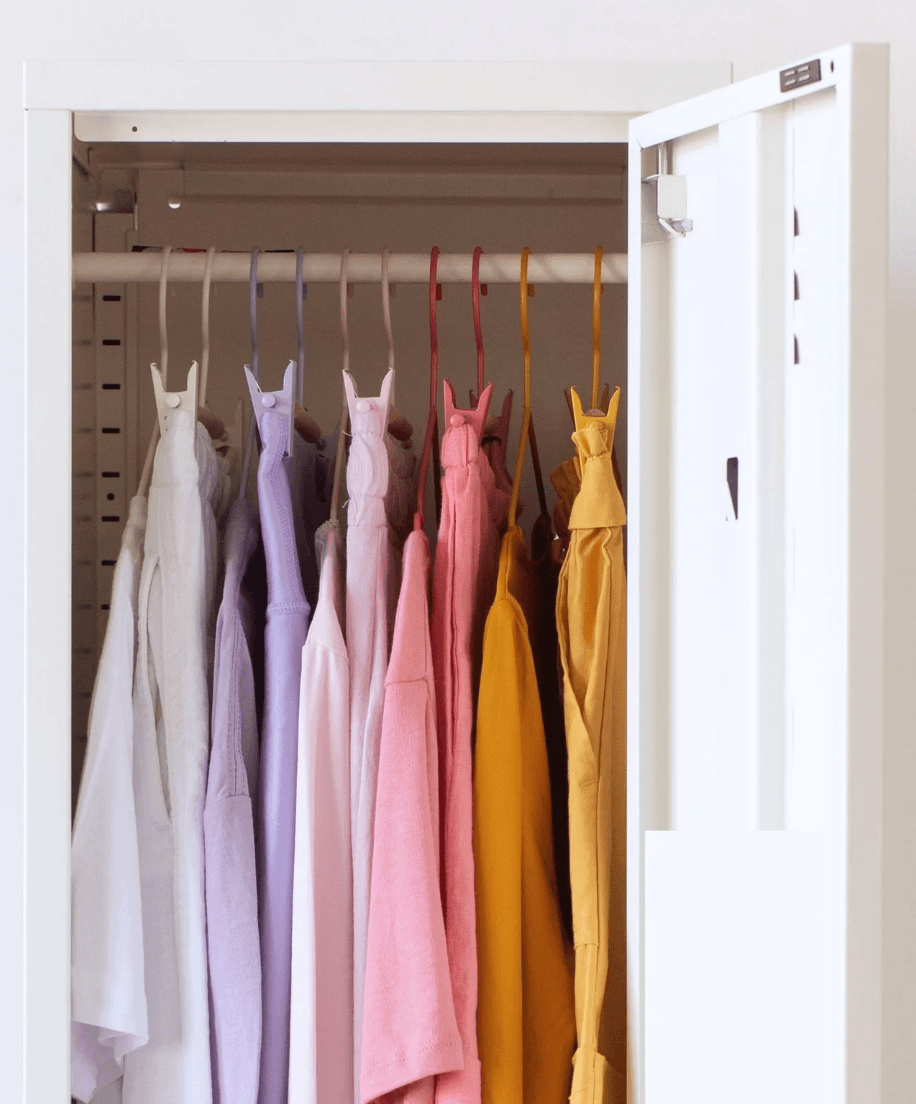 A white wardrobe or closet door is open, showing several pastel and warm-toned garments hanging on hangers inside. The visible clothing includes light gray, lavender, pink, peach, and mustard yellow pieces.