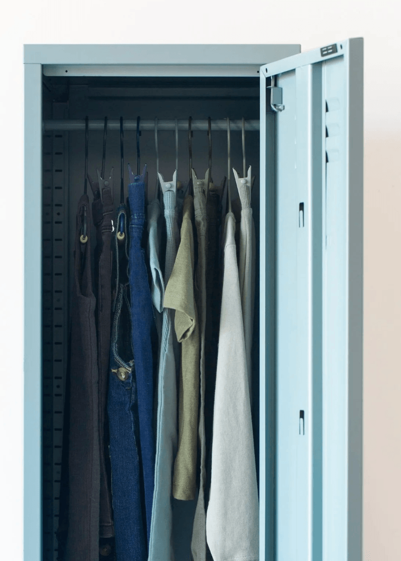 A tall light blue-gray metal garment storage cabinet with its door open, showing hanging clothes inside including jeans, shirts, and a long white garment.
