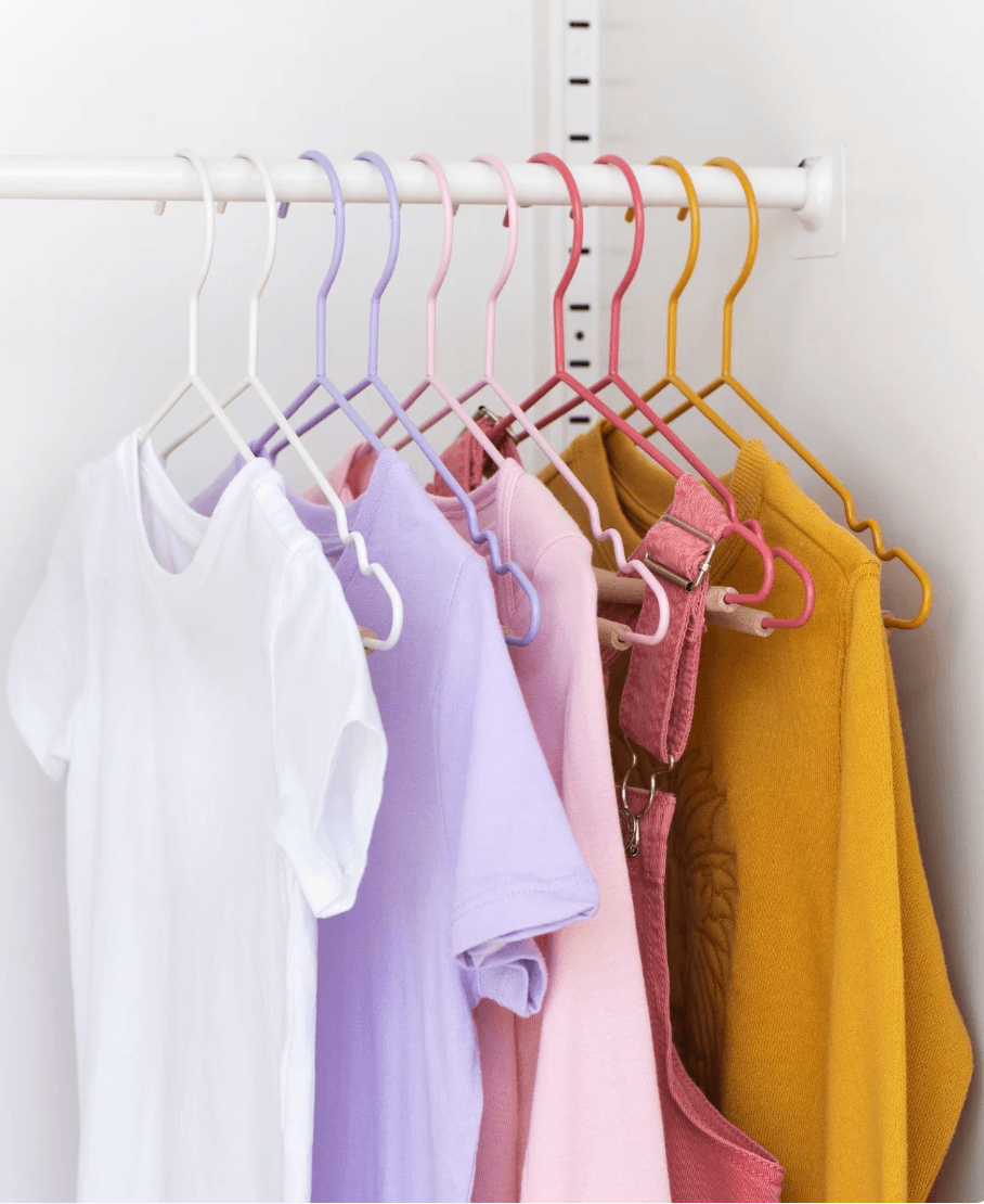 A set of pastel-colored clothes hangers in white, lavender, pink, red, and mustard yellow, holding various colorful garments on a white wall-mounted closet rod.