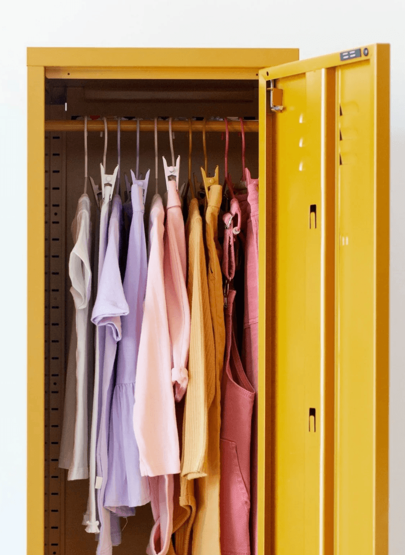 A yellow metal garment storage cabinet with its door open, showing a row of pastel-colored clothing hanging inside on hangers.