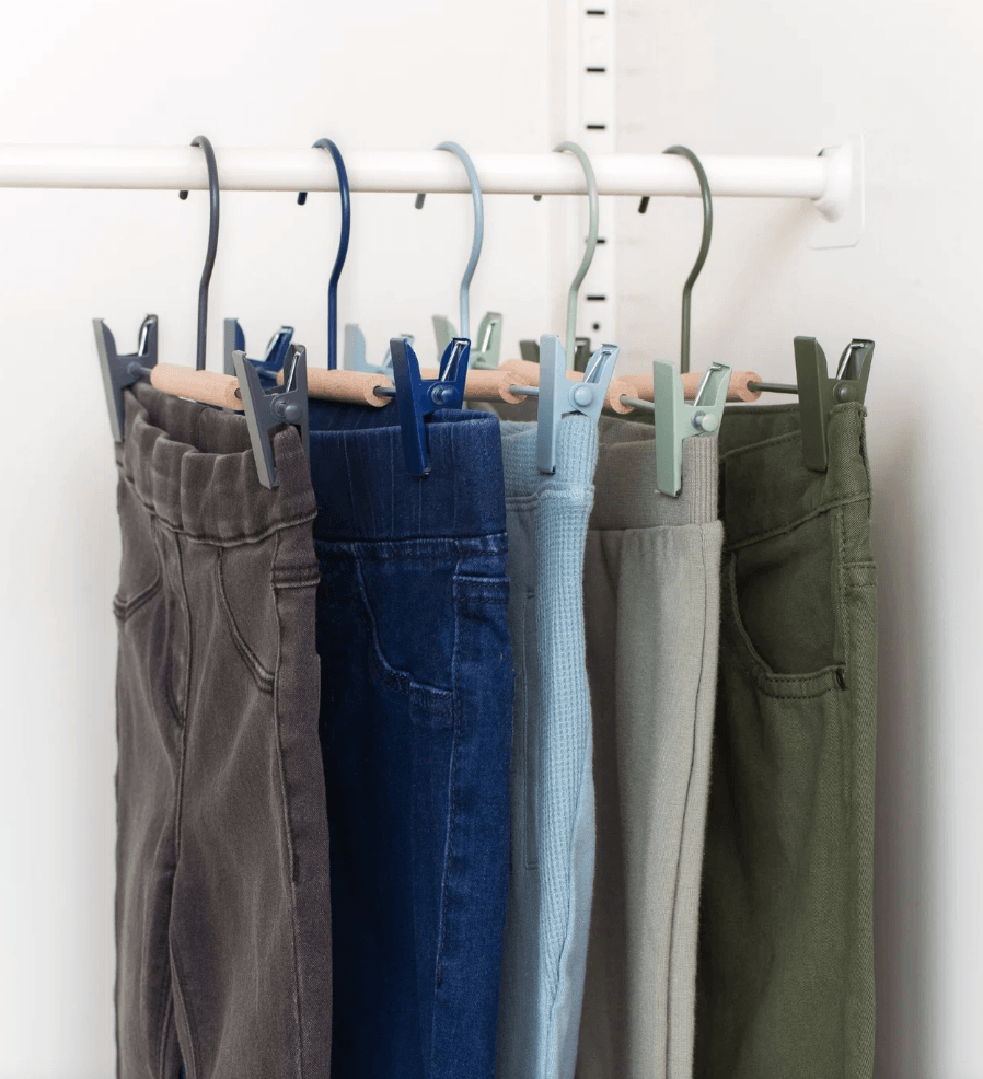 Five pairs of pants hanging from a white closet rod with metal hangers and wooden clothespins, shown against a plain white wall.