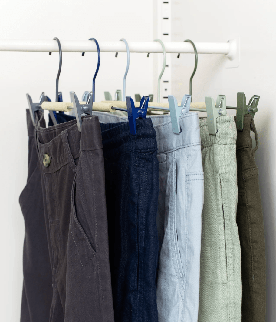 A row of hanging pants in multiple colors—charcoal, navy, light blue, pale green, and olive—secured with blue and green clothespins on a white rail against a white wall.