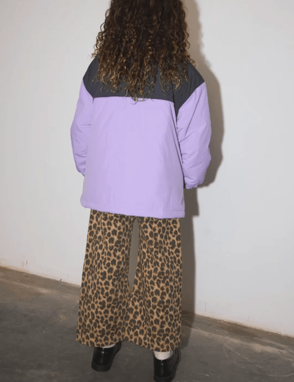 A person wearing a light purple puffer jacket and leopard-print wide-leg trousers, paired with black loafers and white socks, standing against a plain off-white wall.