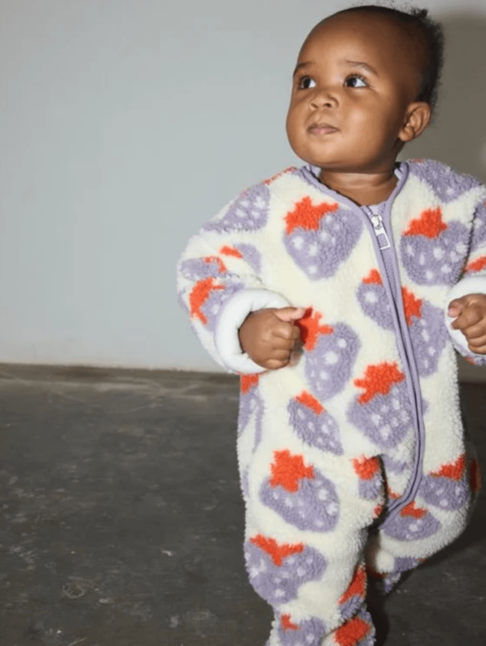 A baby wearing a plush fleece zip-up footed sleeper with a purple and orange strawberry pattern, standing against a plain gray wall.