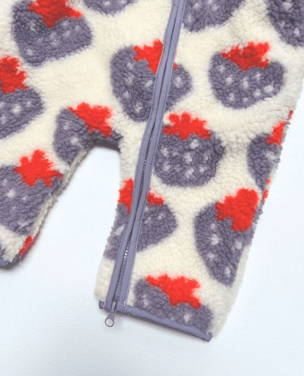 Close-up of a fleece garment with an allover strawberry print on a cream background, featuring purple-gray strawberries with red tops and a matching purple zipper.