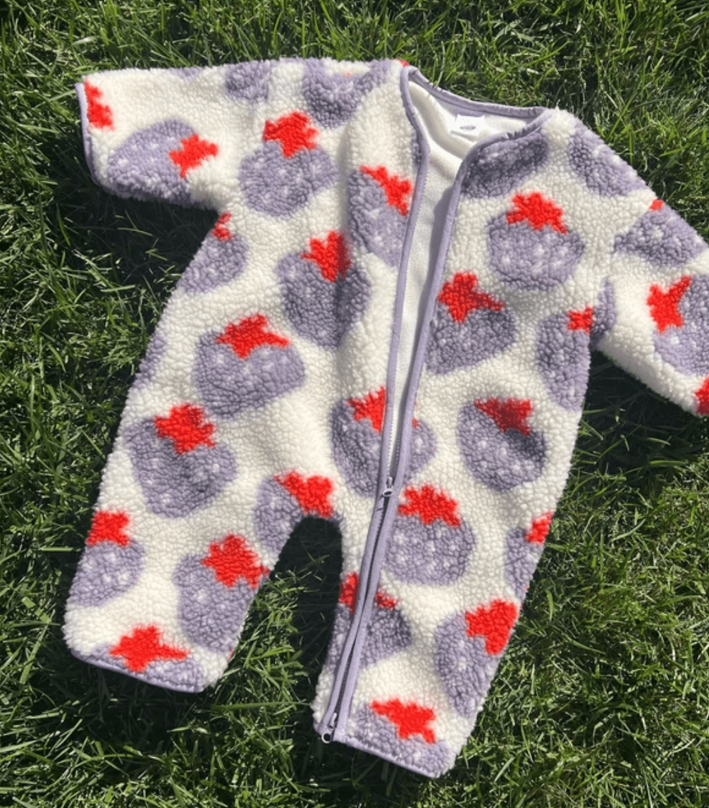 A baby zip-up fleece sleeper laid flat on green grass, featuring a white sherpa-like base with large lavender-purple strawberry motifs and bright red strawberry tops.