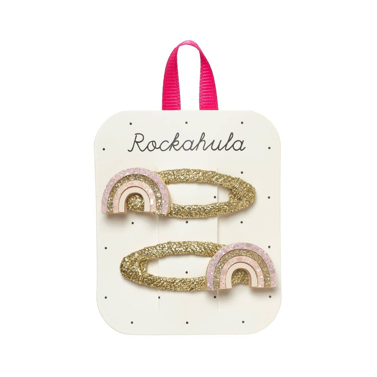 Two gold glitter rainbow hair clips with pink and white accents, attached to a white display card with a pink ribbon loop at the top. The card is printed with the brand name 'Rockahula'.