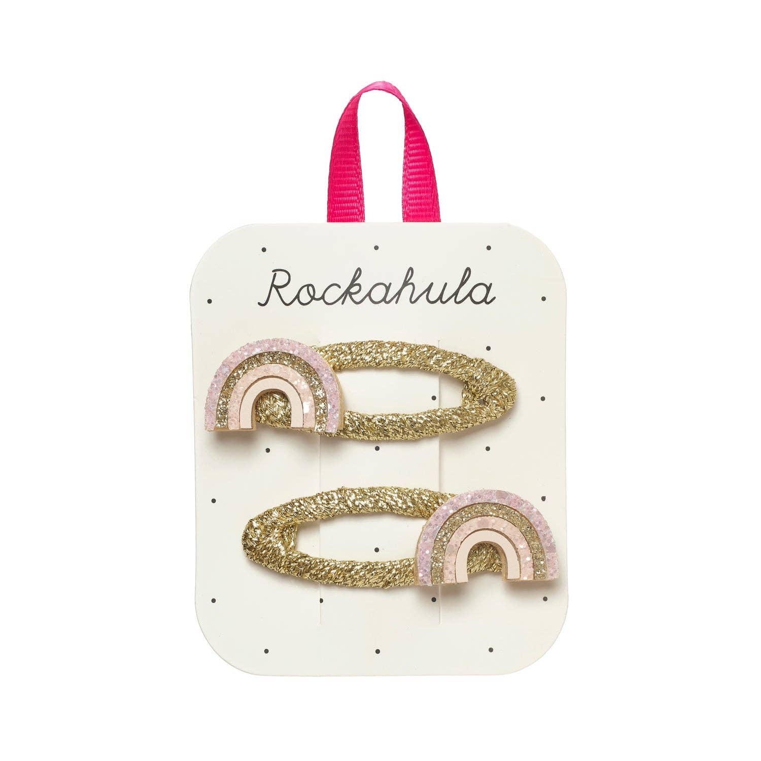 Two gold glitter rainbow hair clips with pink and white accents, attached to a white display card with a pink ribbon loop at the top. The card is printed with the brand name 'Rockahula'.
