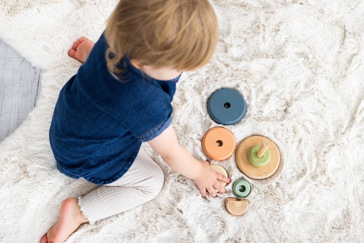 An aerial view of a child wearing a blue shirt sitting on a fluffy carpet playing with a wooden stacking toy consisting of a peg with five multicolor earth tone rings.