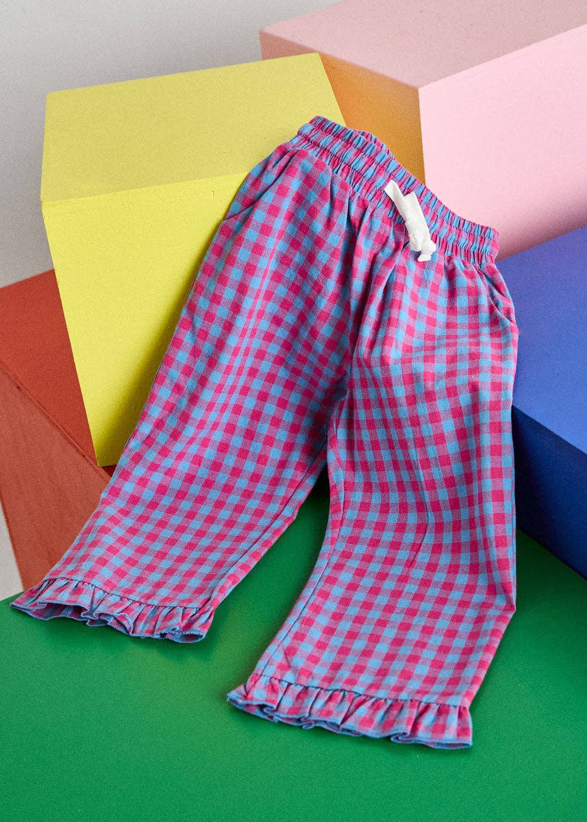 Pink and blue gingham check pajama pants with a ruffled hem, styled on a green surface with colorful geometric blocks in the background.