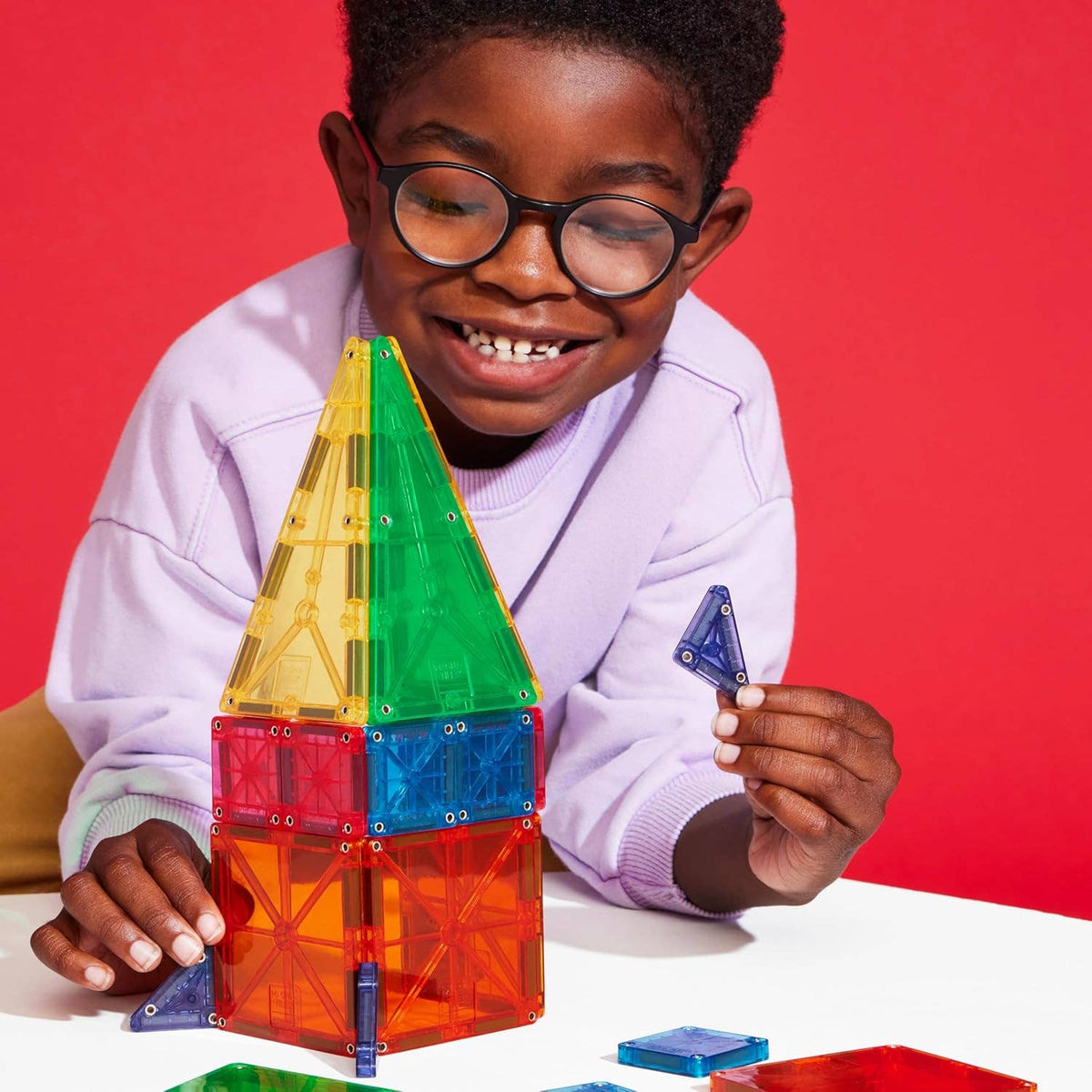 A boy wearing glasses and a lilac sweater is seated at a white tabletop and building a rocket ship with magnetic transparent blocks.