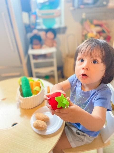 A young child sitting at a wooden table in a bright indoor room, holding a red and green toy fruit. A white plate with two small brown pieces and a basket of colorful toy vegetables are on the table.