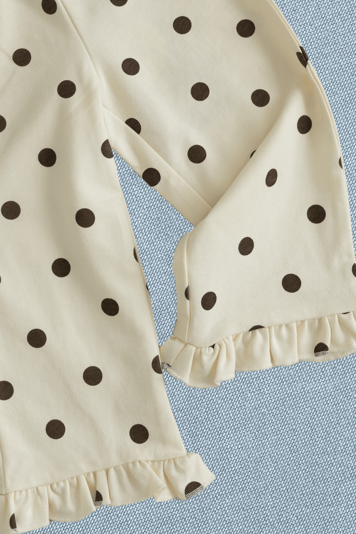 Close-up of a cream-colored garment with dark brown polka dots and ruffled trim, laid flat on a textured light blue fabric background.