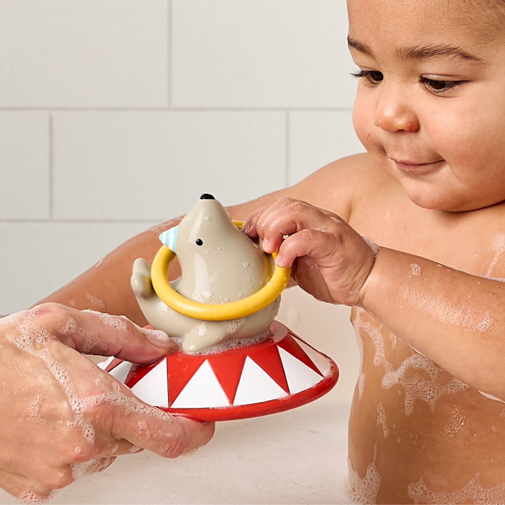 A child in a bubble bath holding a polar bear bath toy that sits on a red and white circus-themed base, with an adult hand supporting it.