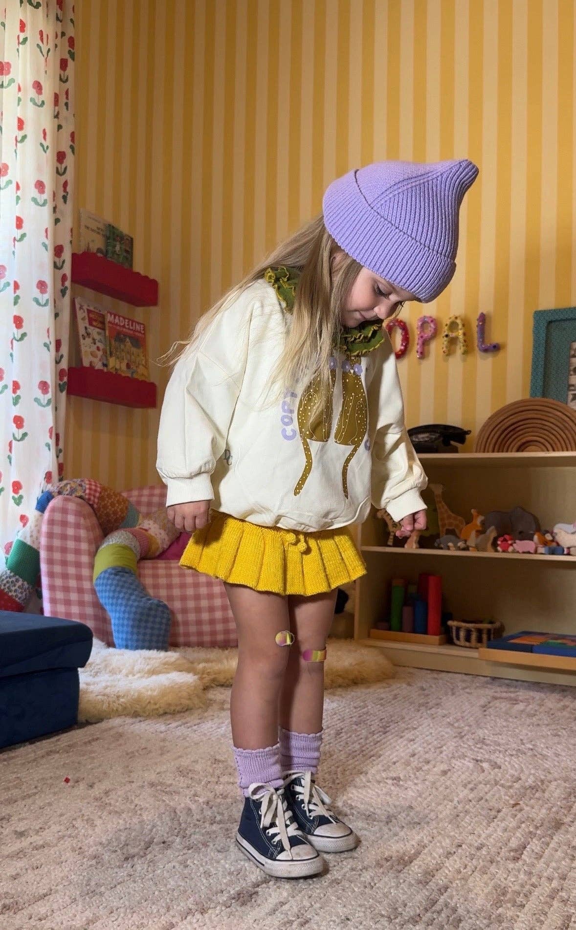 A young child wearing a cream sweatshirt with a gold cat graphic, a yellow pleated skirt, a purple knit beanie, and navy sneakers, standing in a child's playroom.