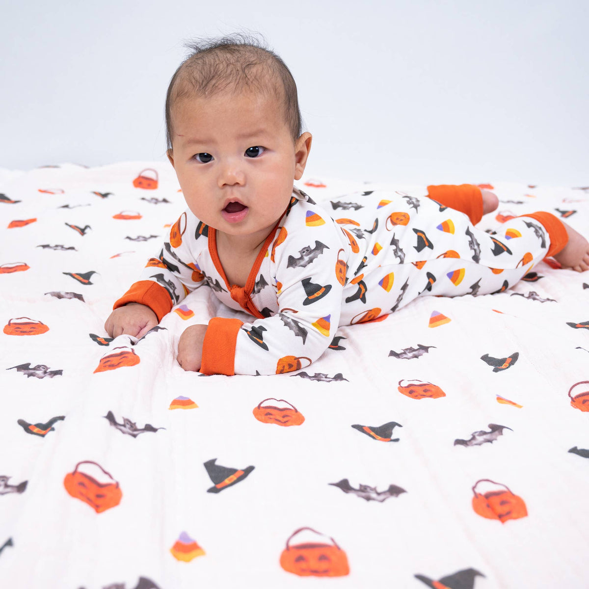 A baby wearing a white one-piece pajama with an orange and black Halloween print, lying on a matching patterned blanket.