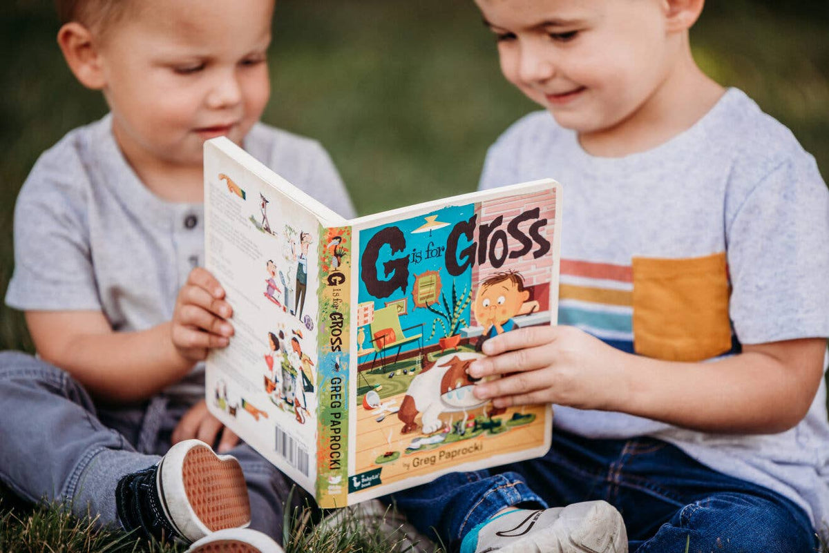 Two children sitting outdoors, reading a book together.