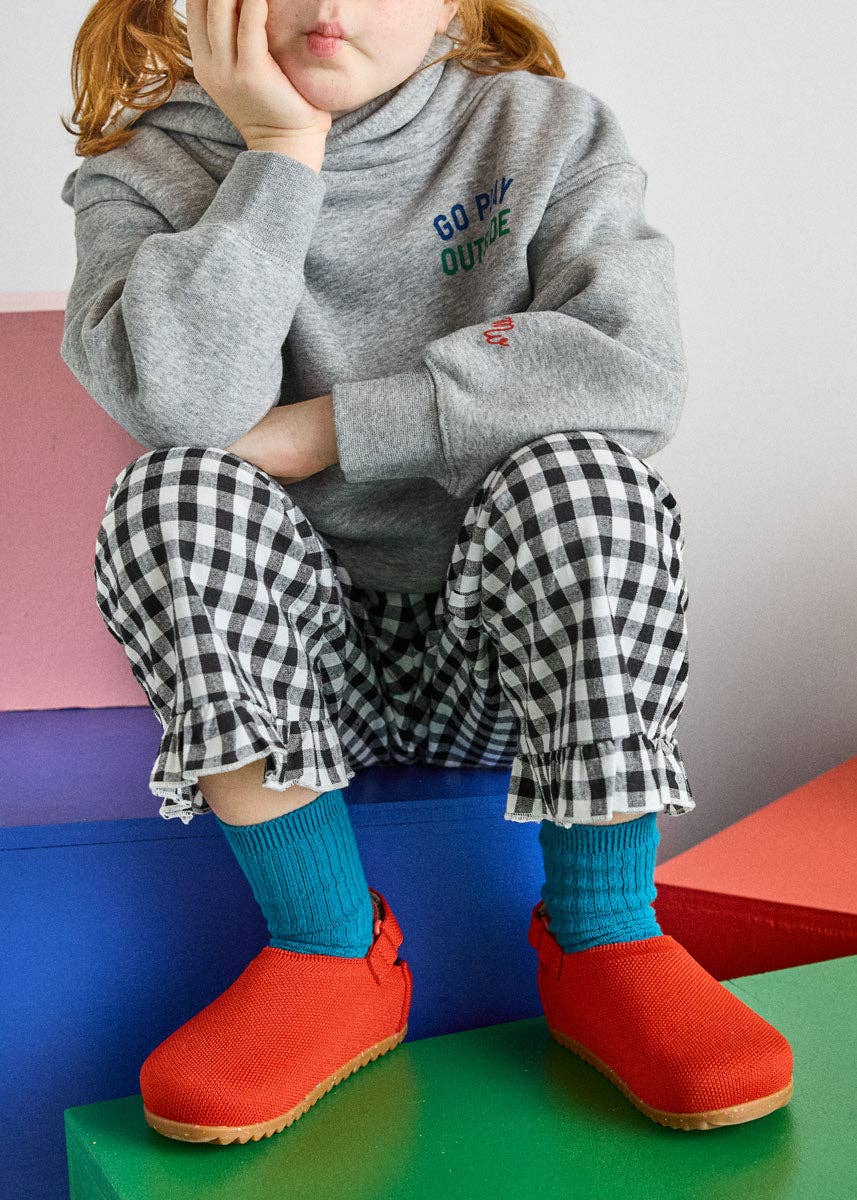 A child wearing a heather gray sweatshirt with colorful text, black and white gingham pants, bright blue socks, and red slip-on shoes, sitting on colorful geometric blocks.