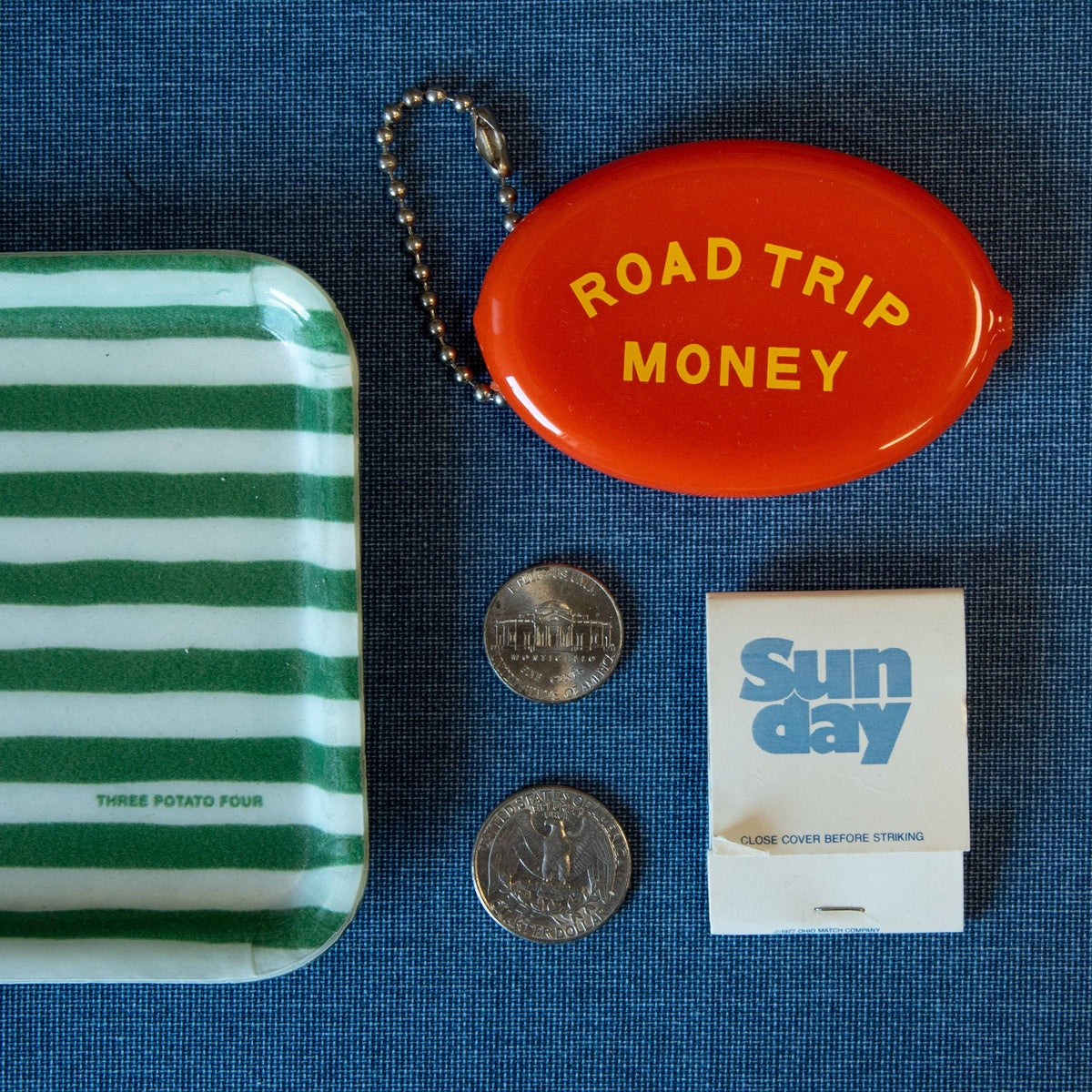 Red container labeled 'Road Trip Money' with coins, a matchbook, and a striped container on a blue textured surface.