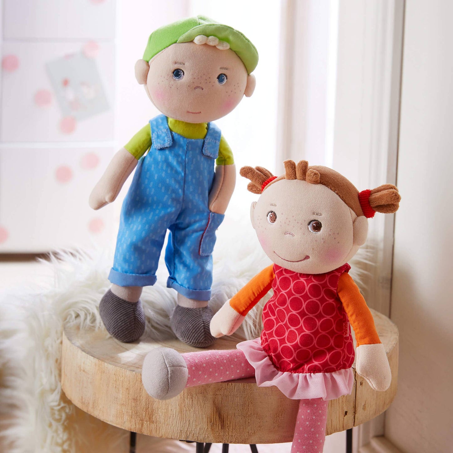 Two dolls, one in blue overalls and green hat, the other in a red dress with pink polka dots, on a wooden stool.