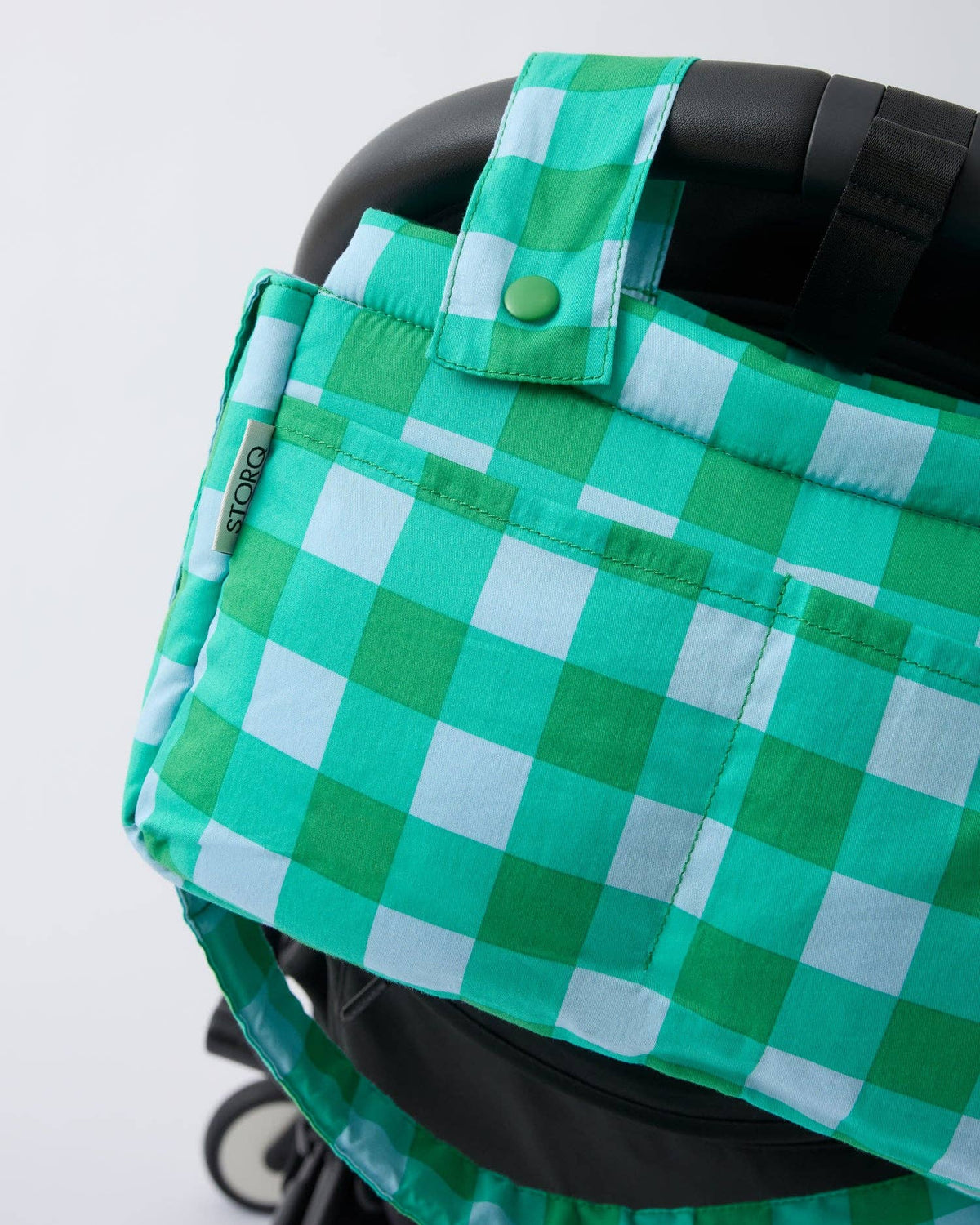 A green and white gingham check stroller cover with a top strap and snap button, shown fitted on a black stroller against a light gray background.
