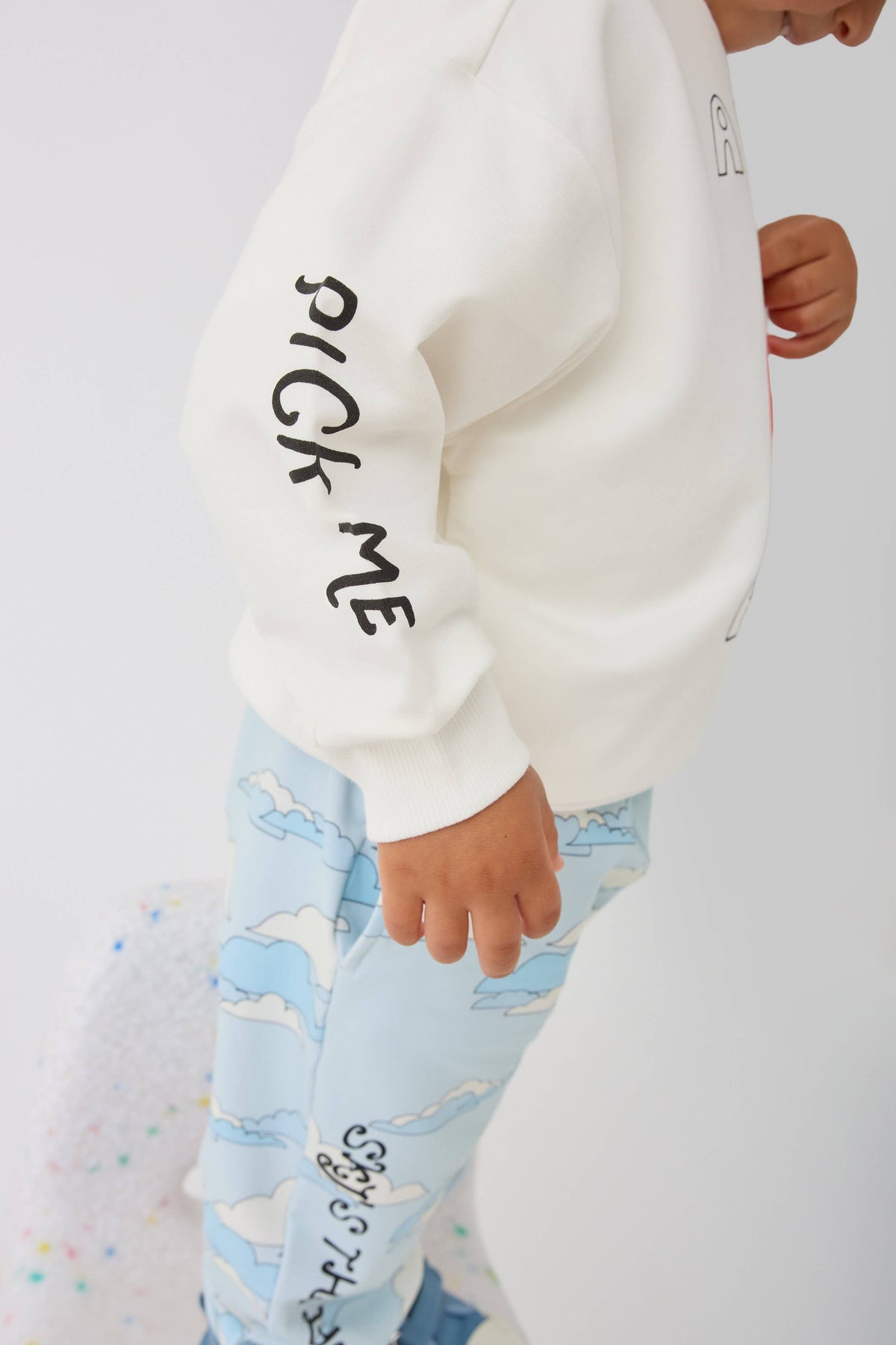 A child wearing a cream sweatshirt with black text on the sleeve and light blue cloud-print pants, shown in a close-up side profile against a white background.