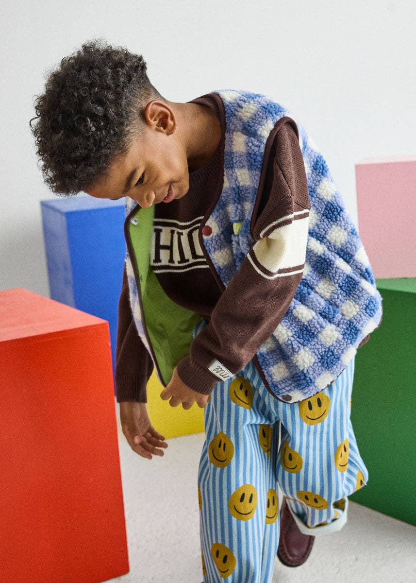 A child wearing a cozy outfit consisting of a blue and white checkered fleece vest, a brown and green graphic shirt, and blue smiley-face patterned pajama pants, standing in front of colorful geometric blocks.