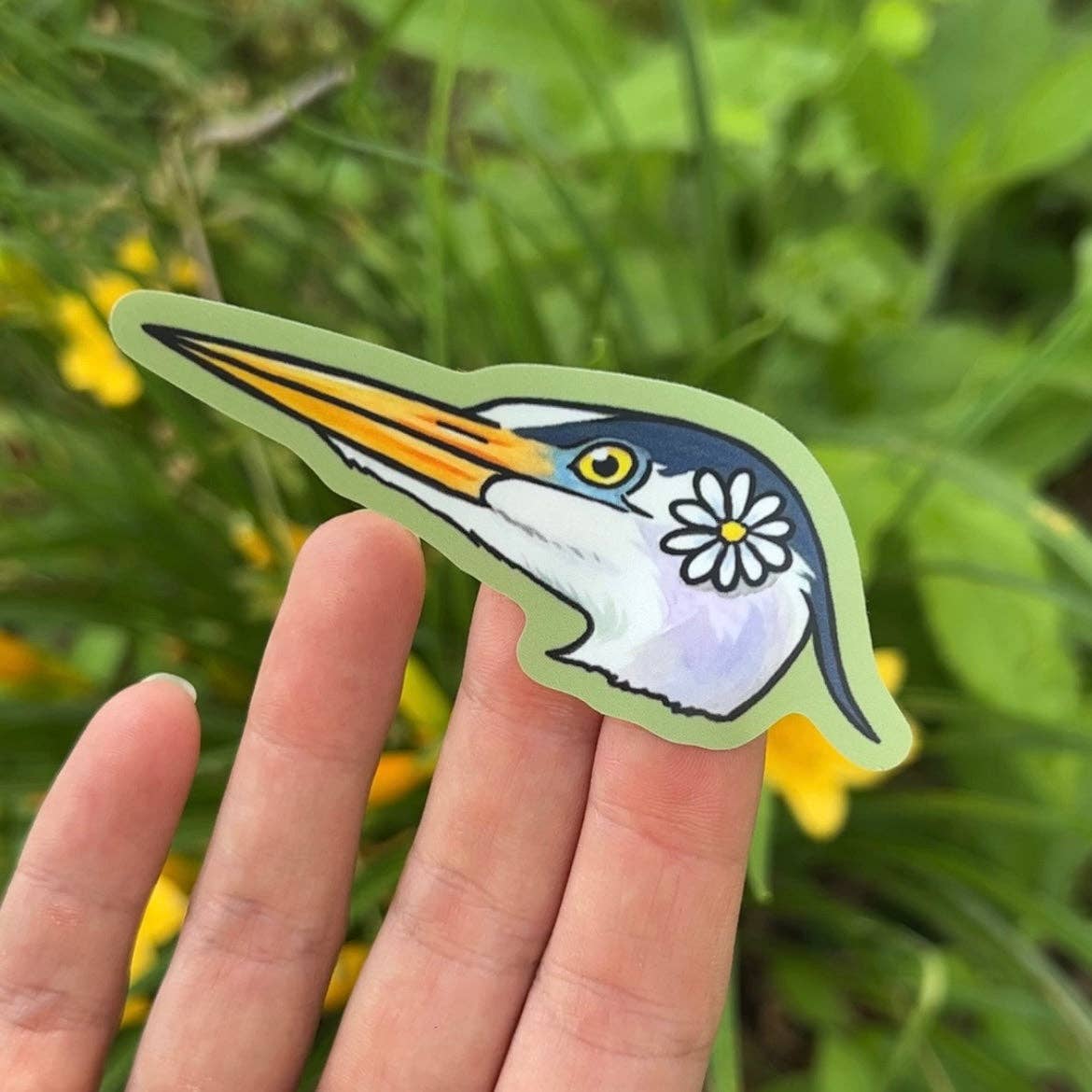 Hand holding a sticker of a heron with a flower on its chest against a natural background.