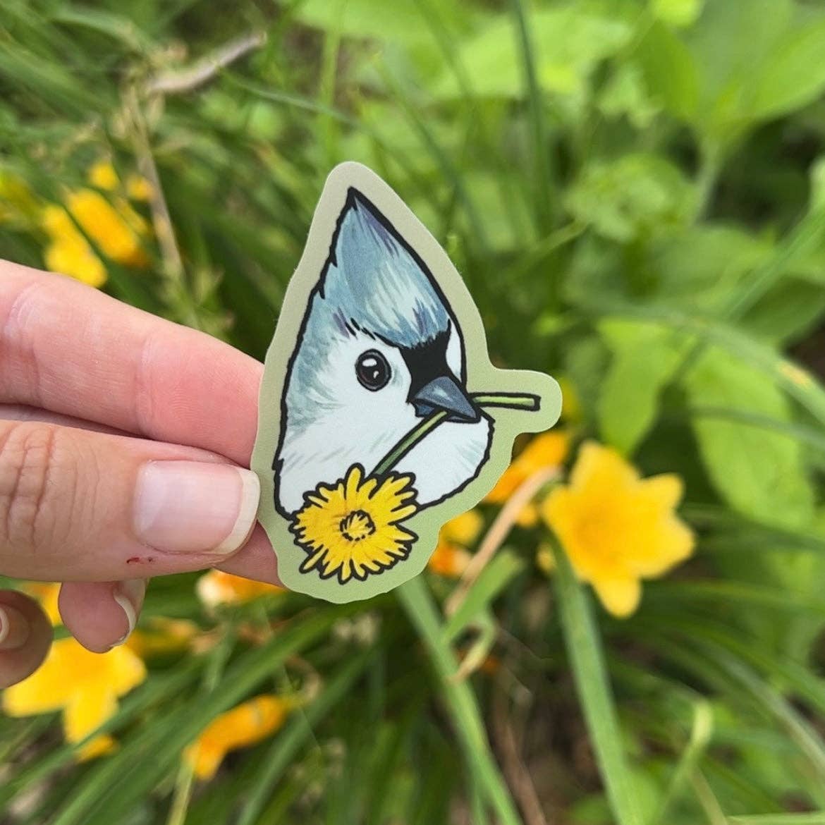 Hand holding a sticker of a bird with a flower in its beak against a natural background with green grass and yellow flowers.