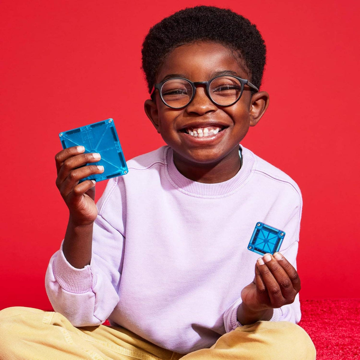 A boy in a lilac sweater is seated against a red backdrop holding two blue transparent sqaure blocks. 
