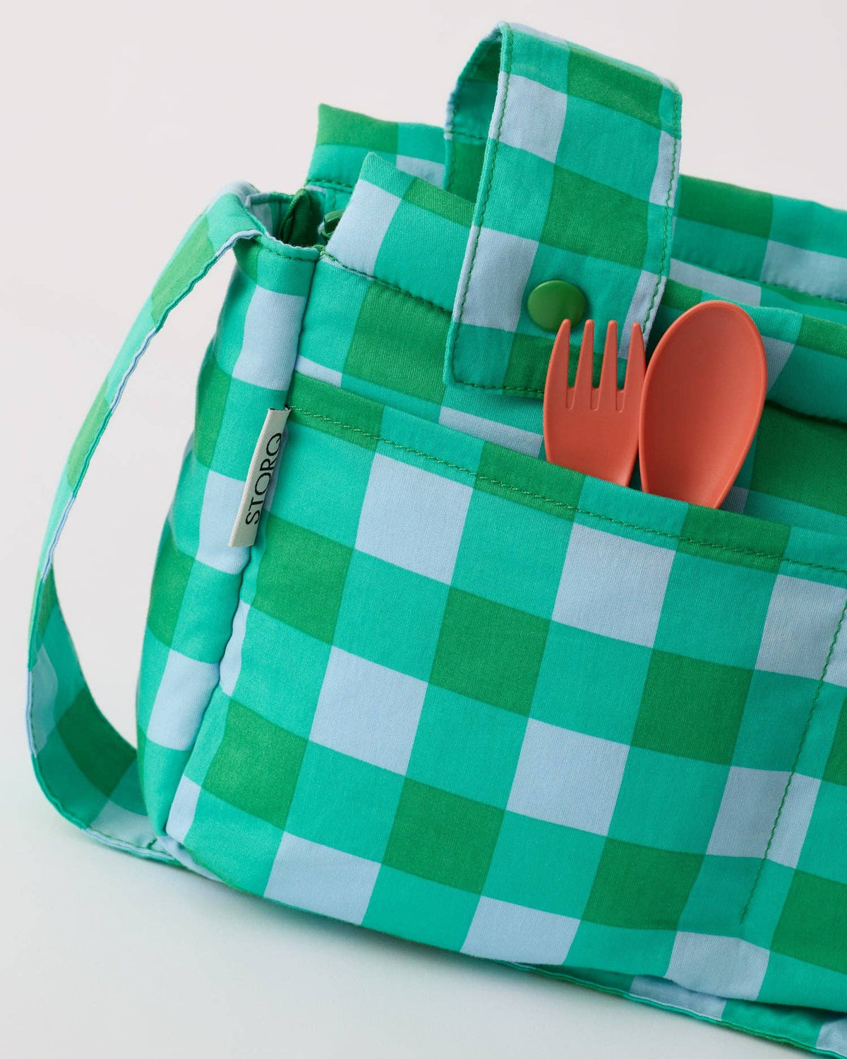 Green and white checkered canvas stroller bag with a shoulder strap, featuring a front pocket holding a green button and coral-colored utensils, with a small fabric tag reading 'storq'.