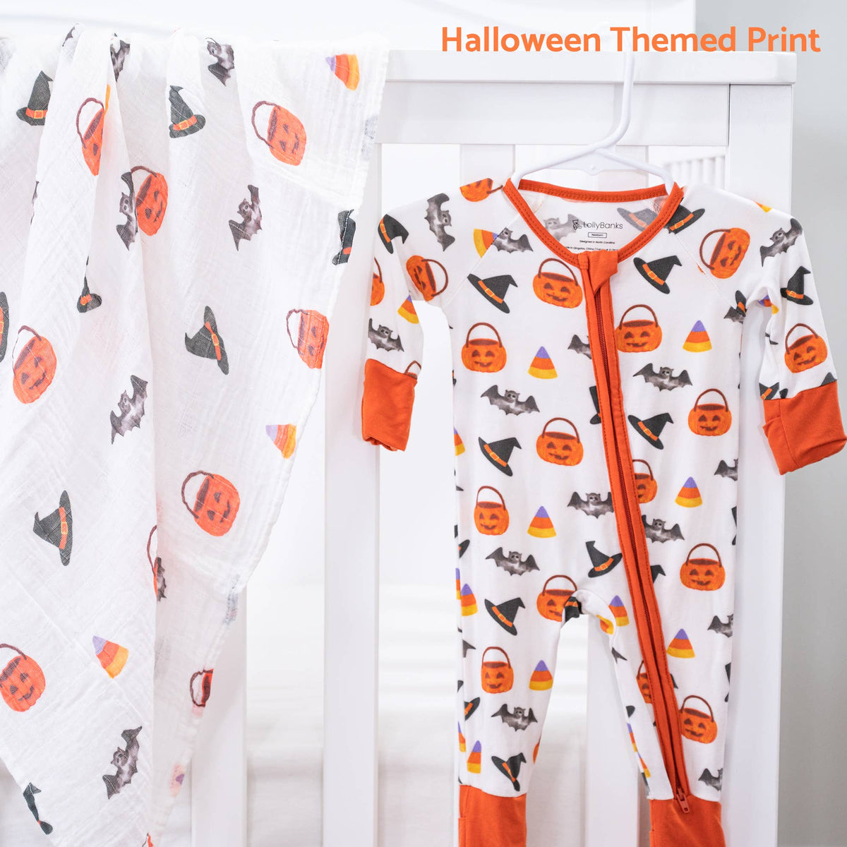 A baby Halloween-themed zip-up footed sleeper in white with orange trim and a print of pumpkins, witch hats, bats, and candy corn, shown hanging next to a matching printed blanket. Text in the top right reads 'Halloween Themed Print'.