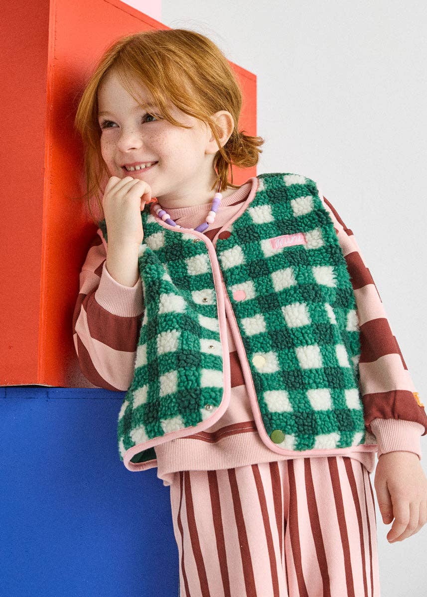 A young girl with red hair smiles while leaning against a colorful geometric wall. She is wearing a green and white checkered fleece vest over a pink and brown striped long-sleeve top, paired with pink and brown striped pants.