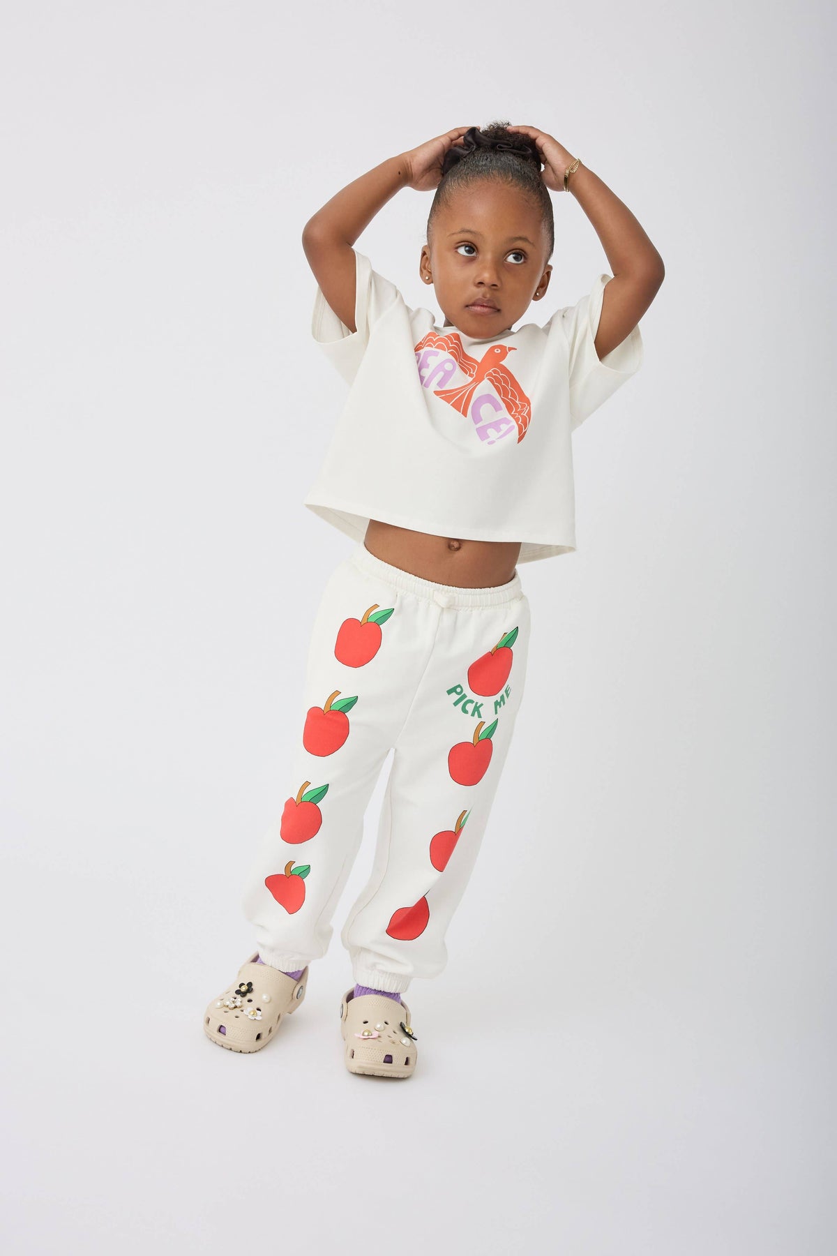 A young child modeling a matching two-piece outfit consisting of a white short-sleeve crop top with a colorful heart graphic and white sweatpants featuring red apple prints and the text 'Pick Me'.