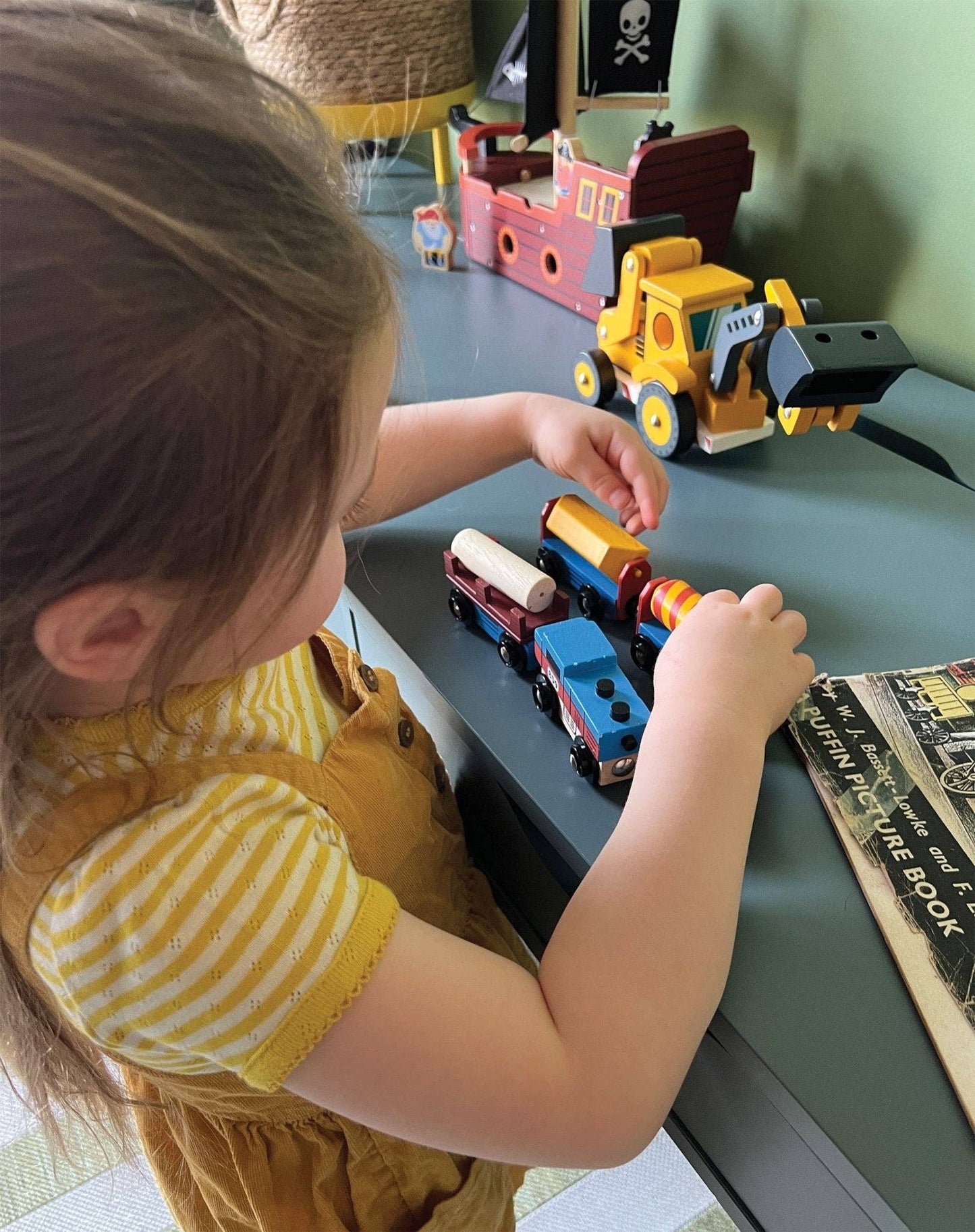 A child wearing a yellow striped sleeve is playing with a wooden train set with one engine and three colorful attached carriages designed to resemble industrial cargo goods.