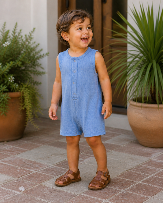 Child wearing a blue romper standing on a patio with plants in the background