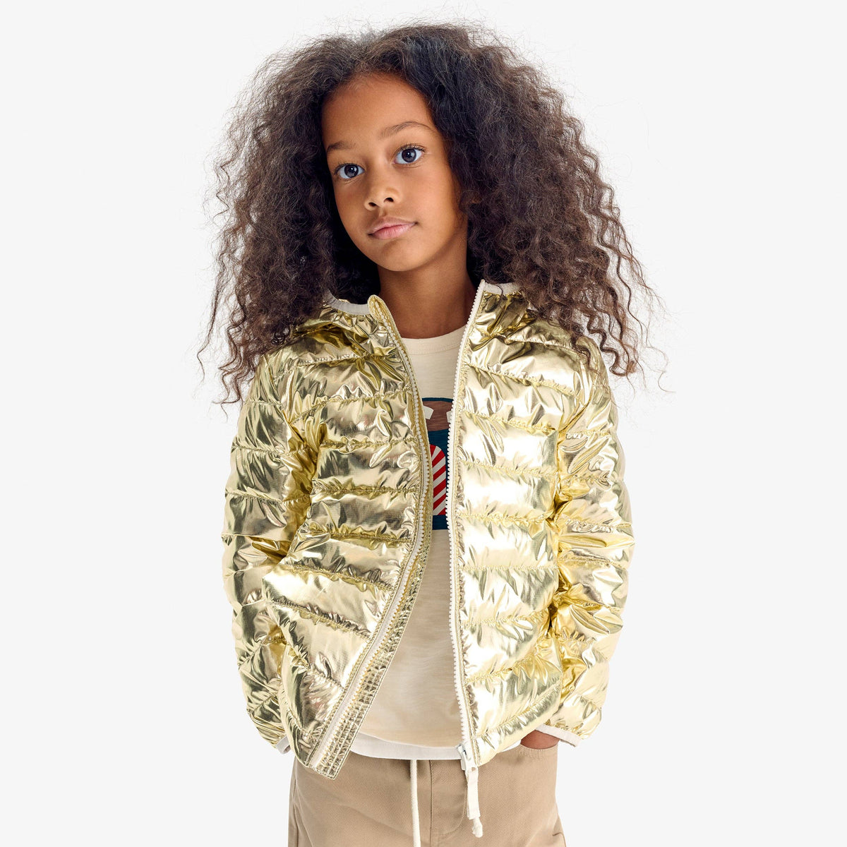 A young girl with long, curly brown hair wearing a shiny metallic gold puffer jacket over a cream-colored t-shirt and beige pants, photographed against a solid light gray background.
