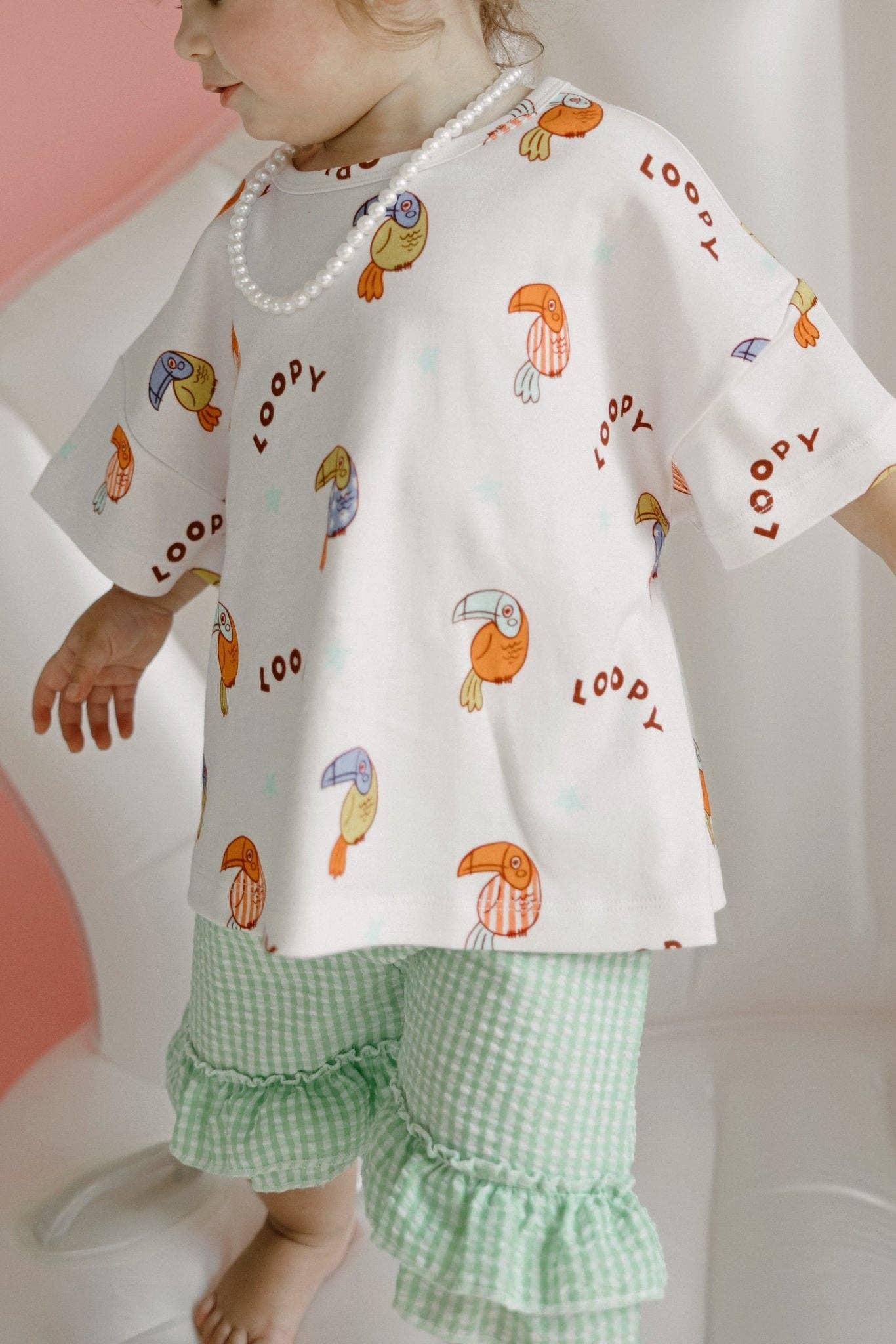 Child wearing a white shirt with colorful bird prints and green checkered pants on a light background