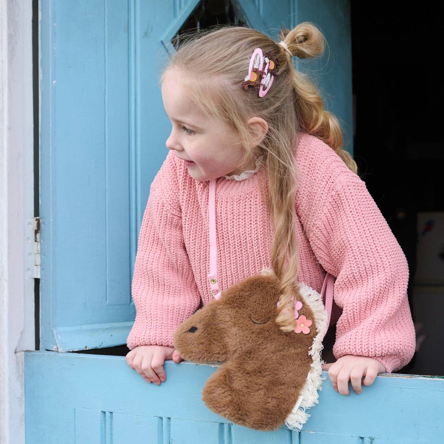 Child in a pink sweater with a horse plush toy, standing in front of a blue door.