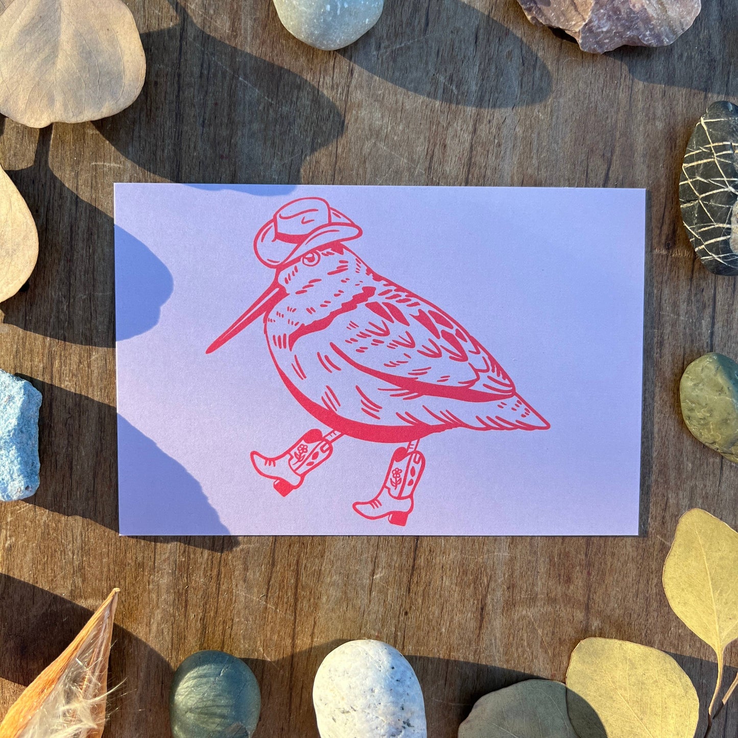 Card with a red bird illustration on a purple background, surrounded by stones and leaves on a wooden surface.
