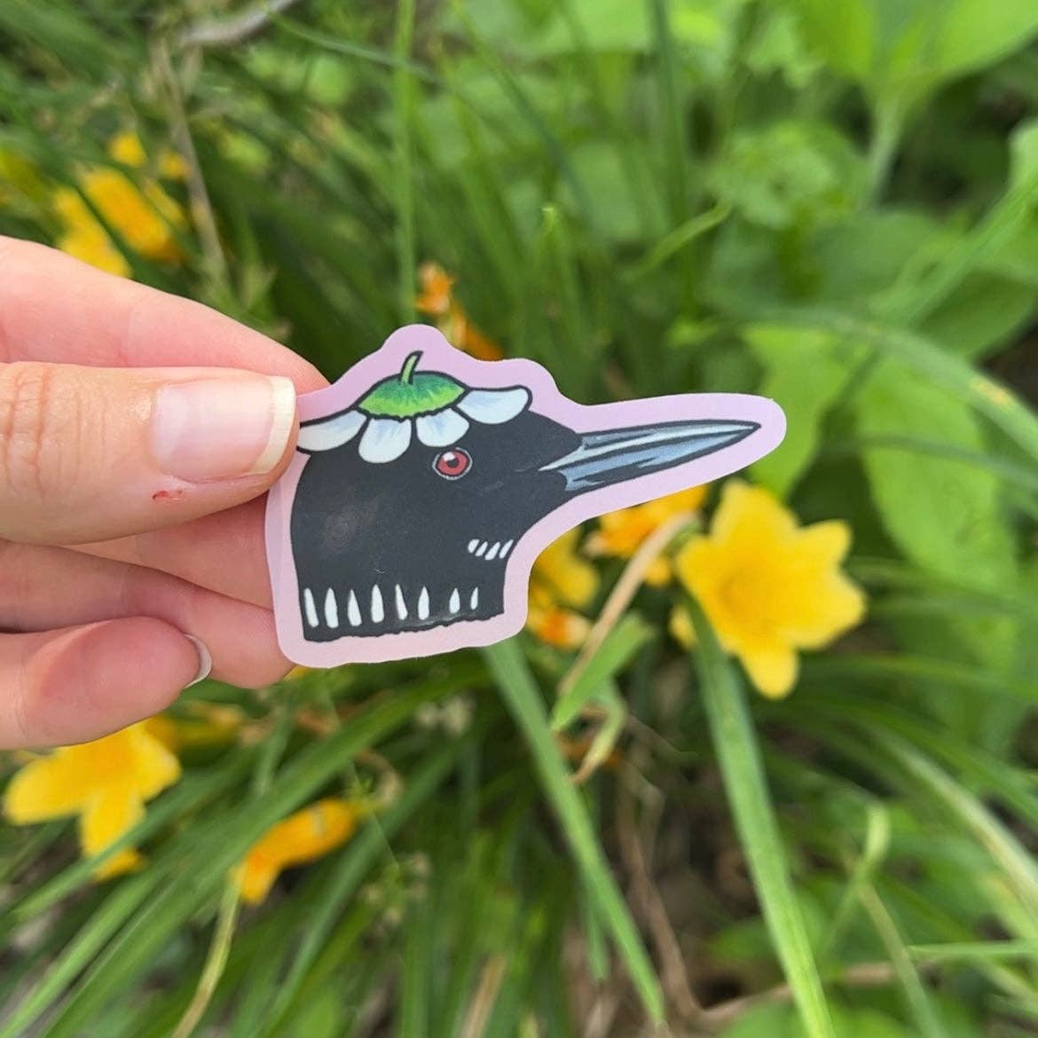 Hand holding a sticker of a cartoonish black bird with a pink border against a natural background with yellow flowers and green grass.