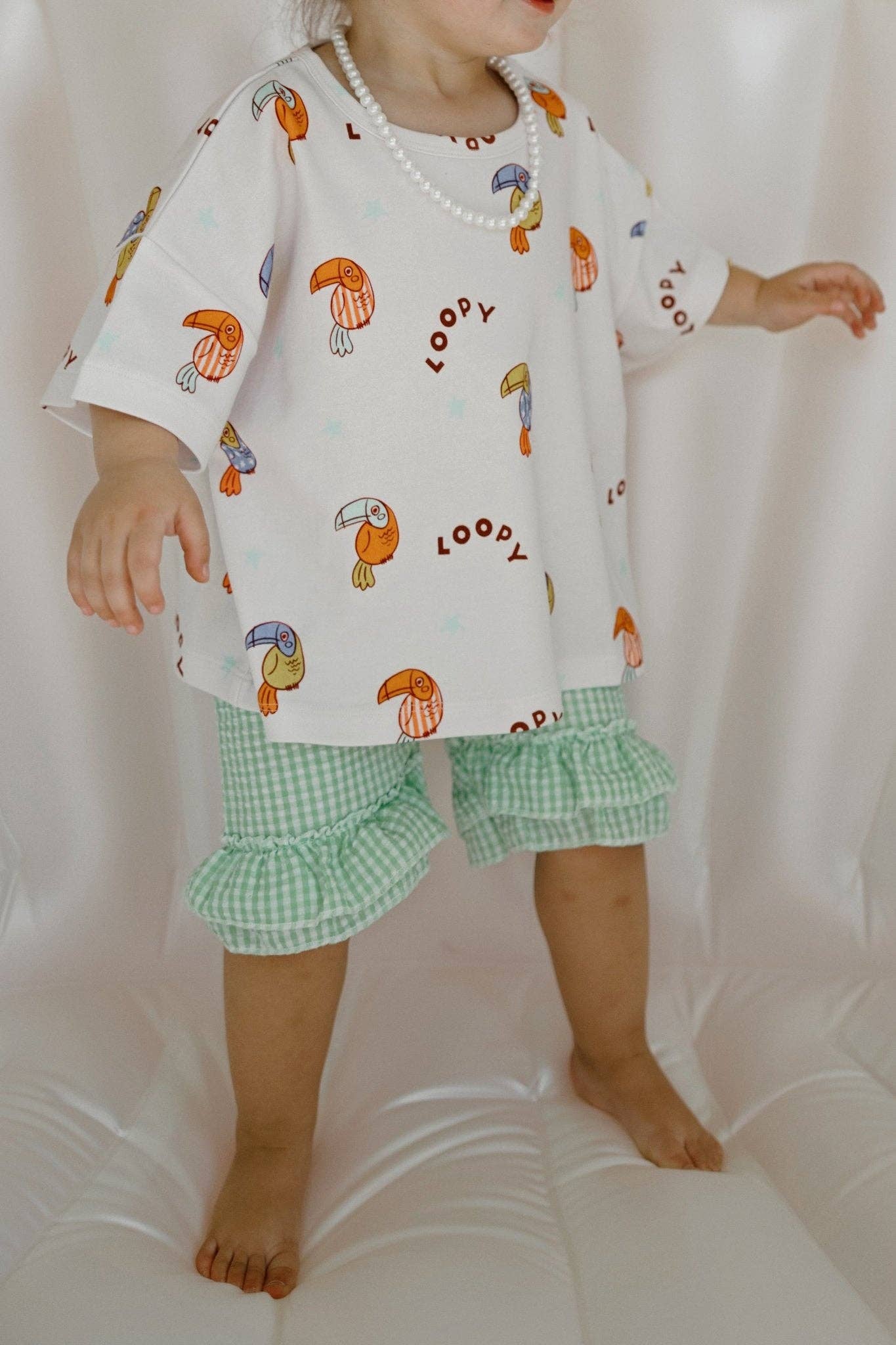 Child wearing a white shirt with toucan prints and green checkered shorts on a white background
