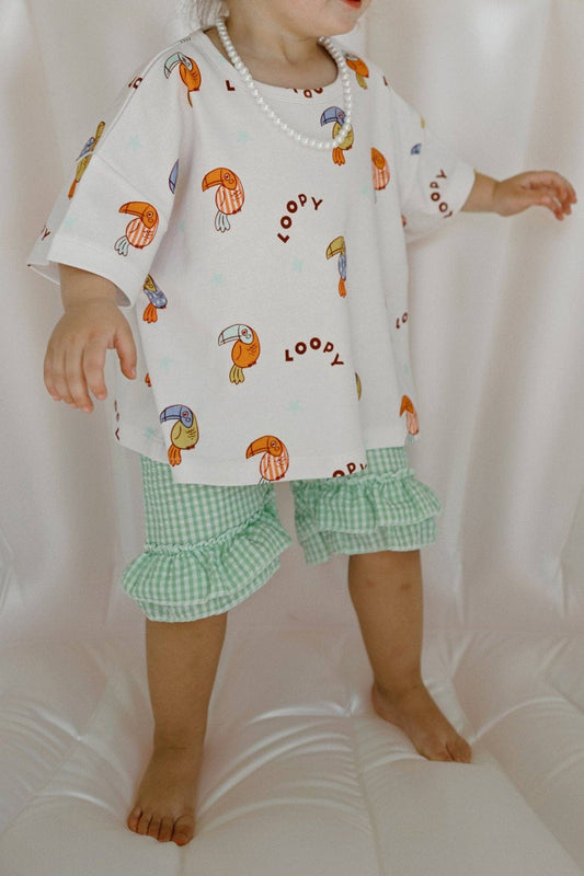 Child wearing a white shirt with toucan prints and green checkered shorts on a white background