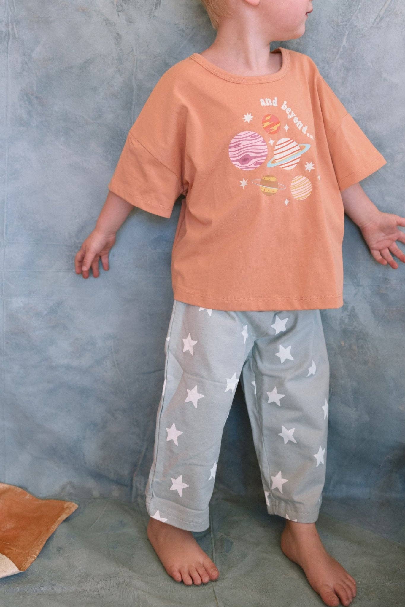 Child wearing an orange t-shirt with a graphic design and light gray pants with star pattern against a gray background