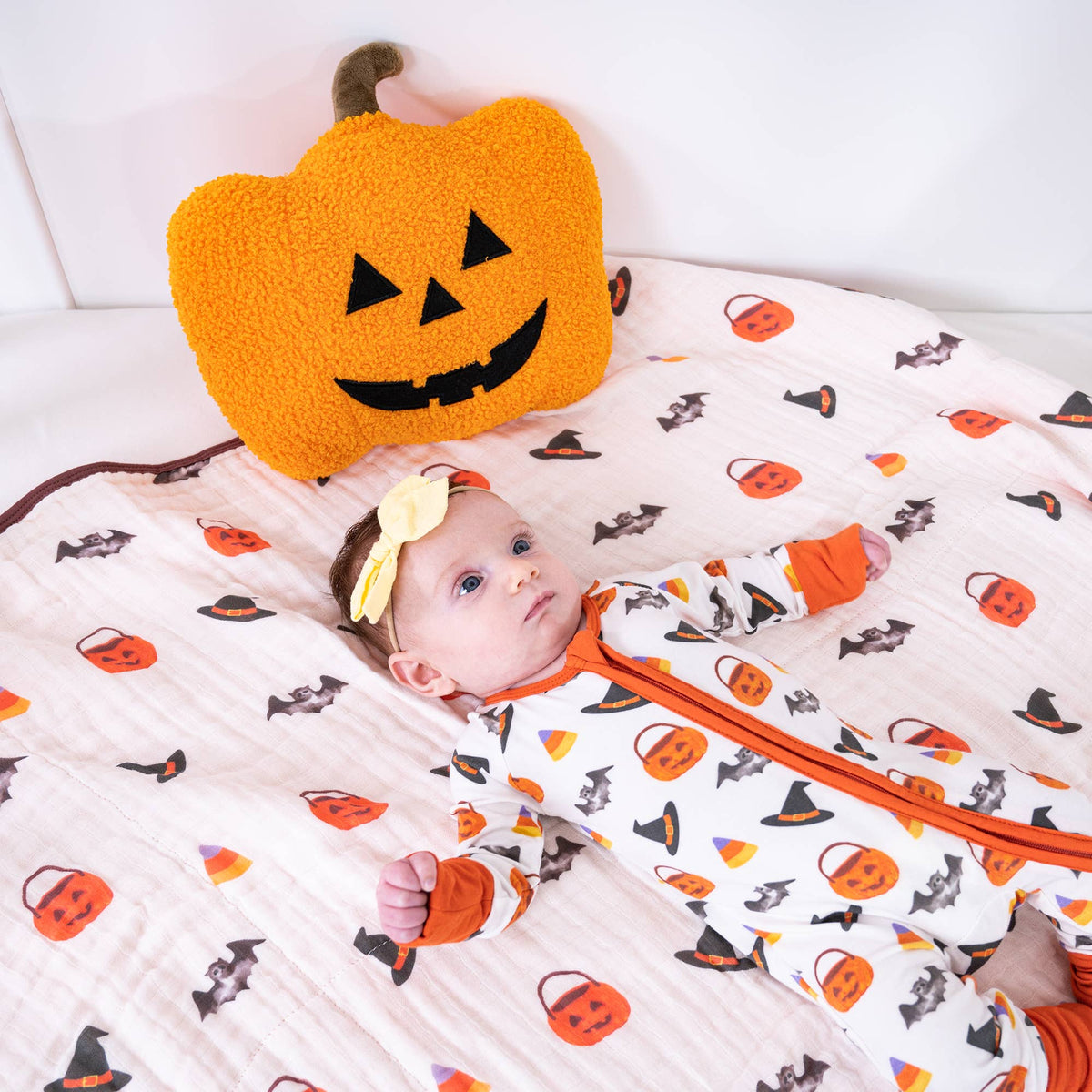 A baby wearing a white zip-up footed sleeper with an orange zipper and an orange, black, and brown pumpkin and witch pattern, lying on a matching patterned blanket next to a plush orange pumpkin pillow.