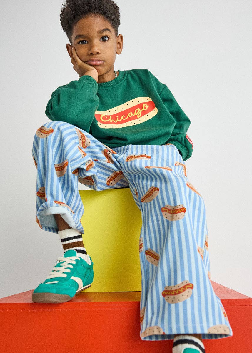 A child model wearing a green sweatshirt with a 'Chicago' graphic and light blue striped pants with a hot dog print, sitting on a yellow block atop a red platform against a light gray background.