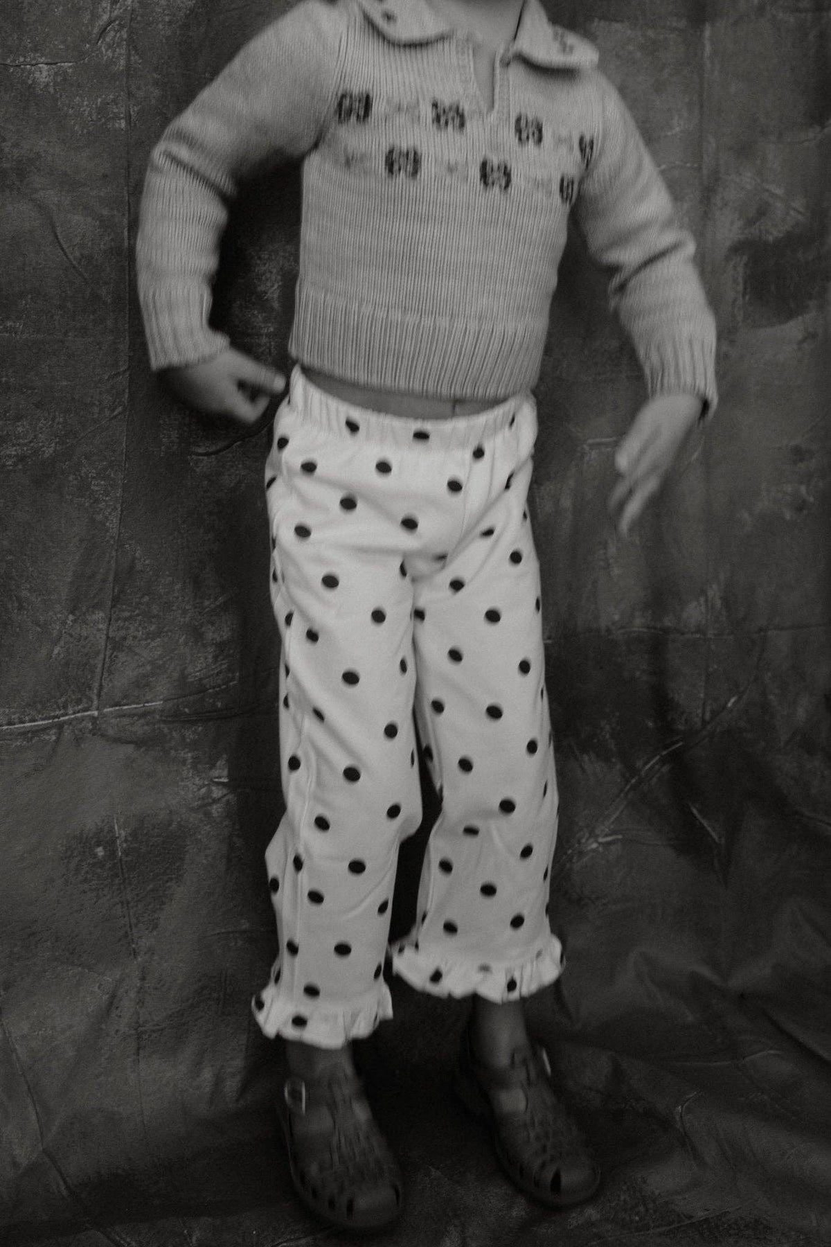 A child wearing a light-colored knit sweater with small motifs and white pants covered in black polka dots, standing against a dark textured backdrop.